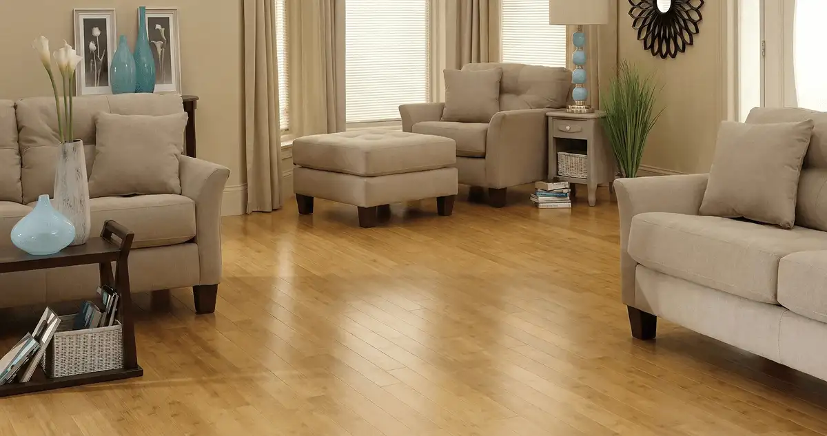 A living room with professionally installed bamboo flooring, beige sofas, a matching ottoman, a side table with a blue lamp, vases, framed pictures, and large windows with blinds and curtains in Pueblo, CO